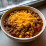 Close-up of a bowl of rich crockpot cowboy soup loaded with ground beef, beans, corn, and topped generously with shredded cheddar cheese.