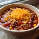 A close-up of a white bowl filled with rich crockpot cowboy soup, topped with shredded cheddar cheese, beans, and corn.