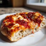 A close-up, appetizing shot of a slice of english muffin pizza showing thick, airy bread base, melted cheese, and rich tomato sauce.