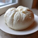 A large, smooth, white ball of freshly made fufu resting on a small white plate near a window.