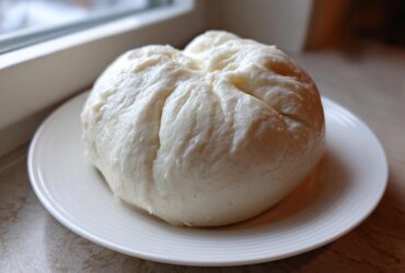 A large, smooth, white ball of freshly made fufu resting on a small white plate near a window.