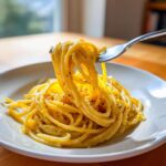 A fork lifting a generous portion of bright yellow Creamy Lemon Pasta, coated in sauce and black pepper.