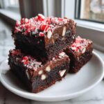Three fudgy candy cane brownies stacked on a white plate, topped with crushed peppermint candy.