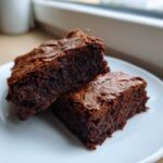 Two rich, fudgy date brownies stacked slightly on a white plate, showing their dense interior.