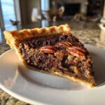 A close-up of a rich, gooey slice of chocolate pecan pie with whole pecans on top, sitting on a white plate.