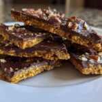 A stack of rich, layered graham cracker toffee pieces topped with melted chocolate and chopped pecans on a white plate.