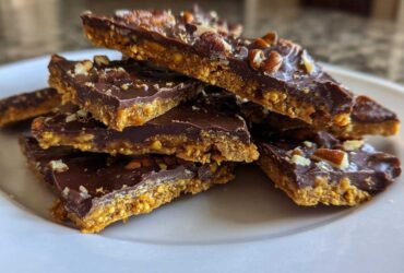A stack of rich, layered graham cracker toffee pieces topped with melted chocolate and chopped pecans on a white plate.