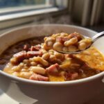 A spoonful of hot ham bean soup, rich with white beans and chunks of ham, lifted from a white bowl.