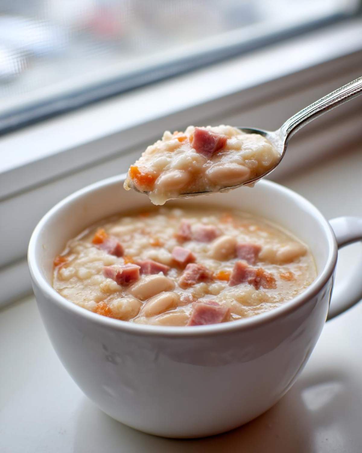 A spoonful of creamy ham bean soup, featuring white beans, diced ham, and carrots, being lifted from a white mug.