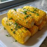 Close-up of several ears of bright yellow corn coated in honey butter and sprinkled with fresh parsley.