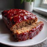 A close-up of a juicy meatloaf loaf with a thick, shiny red glaze, one slice partially cut on a white plate.
