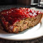A thick slice of juicy meatloaf topped with a thick, glossy red glaze, served on a white plate.
