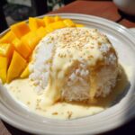 A serving of mango sticky rice, featuring a mound of white rice topped with rich coconut cream and sesame seeds, served alongside fresh mango slices.