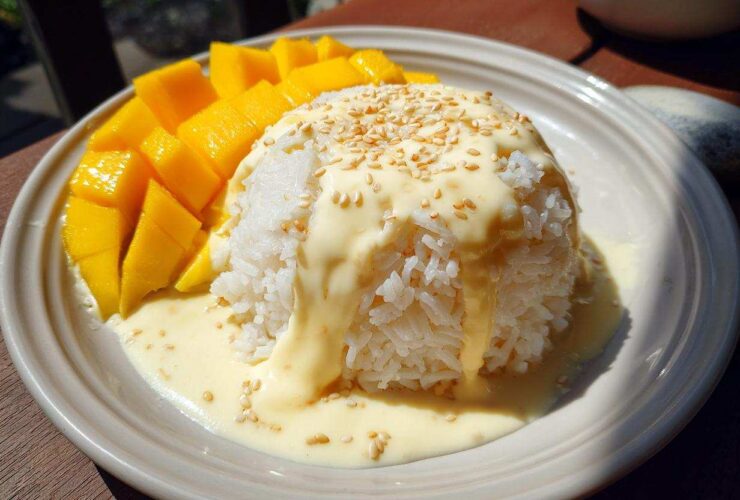 A serving of mango sticky rice, featuring a mound of white rice topped with rich coconut cream and sesame seeds, served alongside fresh mango slices.
