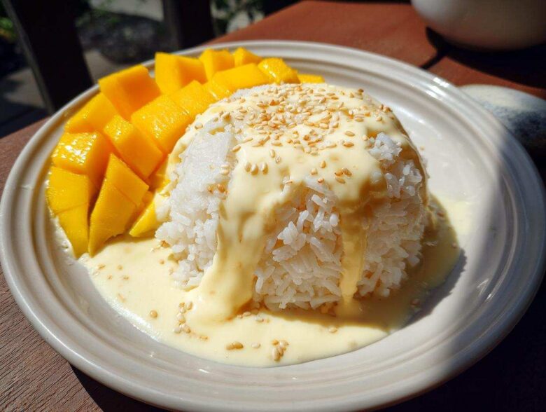 A serving of mango sticky rice, featuring a mound of white rice topped with rich coconut cream and sesame seeds, served alongside fresh mango slices.