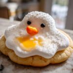 A close-up of one melted snowman cookies featuring white frosting, black eyes, an orange carrot nose, and yellow 'puddle' icing.