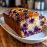 A freshly baked, golden brown blueberry banana bread loaf studded with juicy blueberries, resting on a white platter.