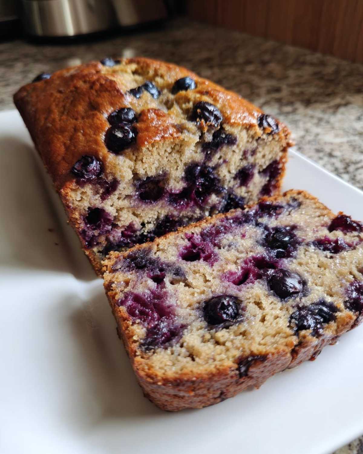 Close-up of a moist blueberry banana bread loaf, showing a thick slice revealing juicy blueberries throughout the crumb.