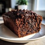 A close-up of a rich, dark chocolate banana bread loaf topped with melted chocolate chips, sitting on a white plate.