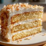 A thick slice of moist, three-layer coconut cake frosted with white icing and covered in toasted coconut flakes.