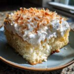 A close-up of a moist slice of coconut poke cake topped with whipped cream and toasted coconut flakes.