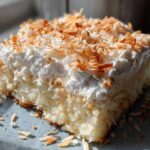A close-up of a moist slice of coconut poke cake topped with whipped cream and toasted coconut flakes.