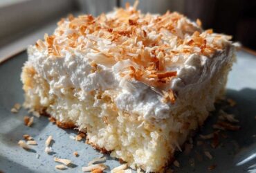 A close-up of a moist slice of coconut poke cake topped with whipped cream and toasted coconut flakes.
