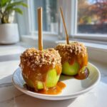 Two bright green caramel apples dipped in thick caramel sauce and covered with chopped nuts, served on a white plate.