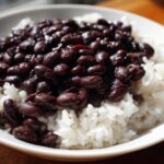 A close-up of a white bowl filled with fluffy white rice topped generously with saucy black beans, perfect for a beans and rice dinner.