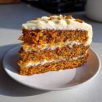 A perfect slice of layered carrot cake recipe showing moist crumb, cream cheese frosting, and chopped nuts on top.