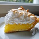 Close-up of a tall slice of lemon meringue pie showing bright yellow filling and toasted meringue topping.