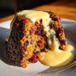 Close-up of a dense slice of plum pudding loaded with dried fruits, drizzled generously with creamy yellow custard.