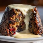A close-up of a dark, moist plum pudding, cut open to show dried fruit, topped with thick vanilla sauce.