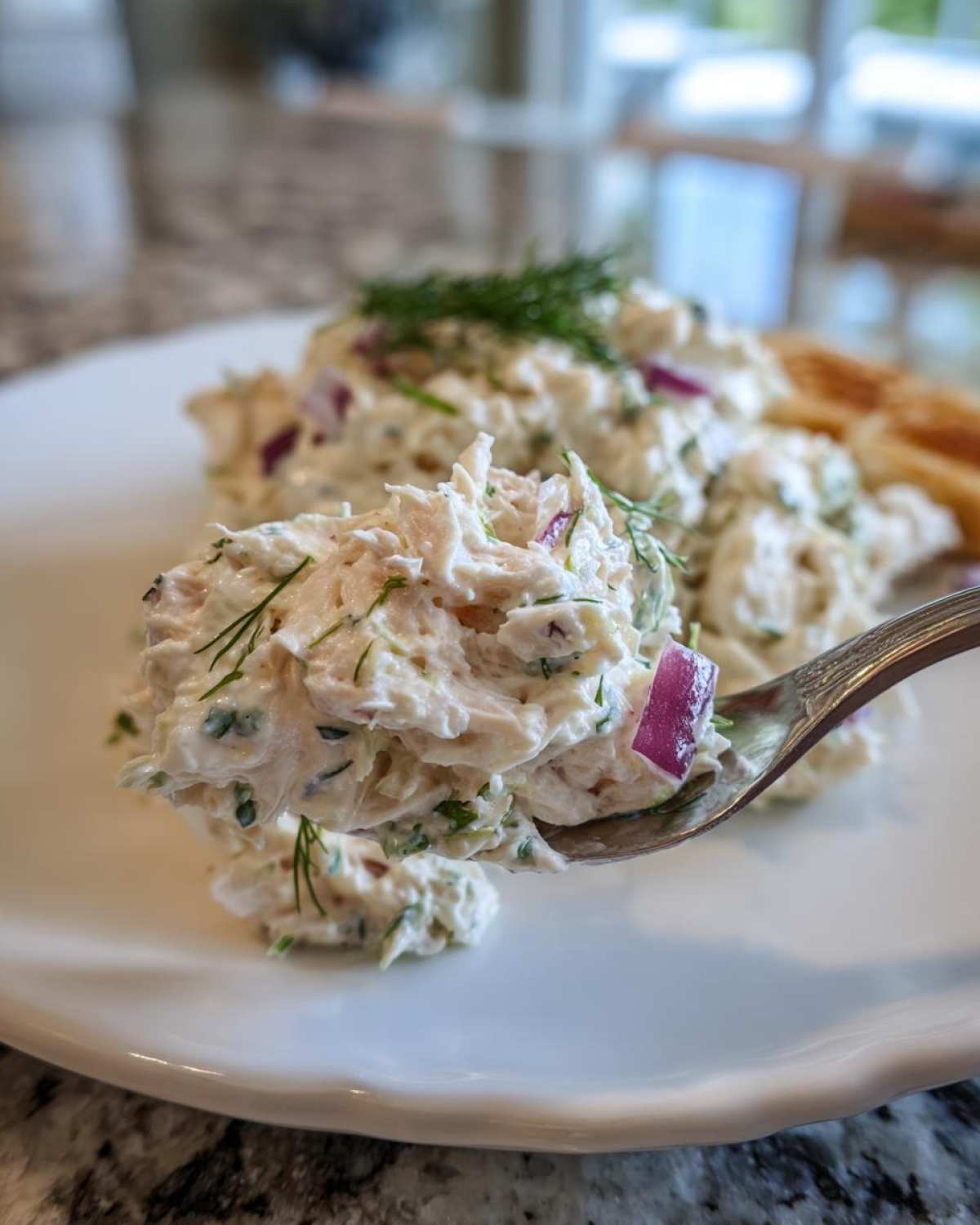 A spoonful of creamy cottage cheese chicken salad mixed with red onion and fresh dill being lifted from a white plate.