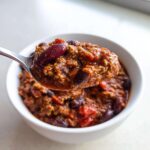 A spoonful of thick, rich high protein chili, featuring ground meat and dark red kidney beans, lifted from a white bowl.