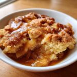 Close-up of a warm serving of apple cobbler in a white bowl, topped with rich caramel sauce.