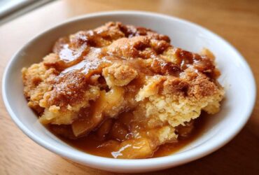 Close-up of a warm serving of apple cobbler in a white bowl, topped with rich caramel sauce.
