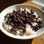 A white bowl filled with fluffy white rice topped generously with dark black beans, perfect for a quick beans and rice dinner.