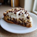 A thick slice of chocolate chip cookie cake recipe topped with white frosting and mini chocolate chips.
