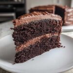 Close-up of a moist slice of homemade chocolate cake layered with rich chocolate frosting on a white plate.