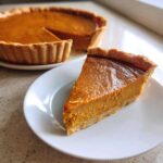A perfect slice of easy pumpkin pie resting on a white plate, with the rest of the pie visible in the background.
