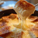 A piece of toasted bread is lifted from a pot of smooth cheese fondue, showing long, gooey cheese pulls.
