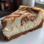 A close-up of a creamy slice of snickerdoodle cheesecake with a cinnamon swirl topping and graham cracker crust.