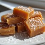 Close-up of soft, chewy caramels sprinkled with flaky sea salt, piled on a light-colored plate.
