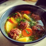 Close-up of a steaming bowl of albondigas soup, featuring browned meatballs, chunks of potato, sliced carrots, and fresh cilantro.