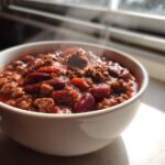 Close-up of a steaming white bowl filled with hearty chili, featuring ground meat and red kidney beans.