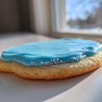 Close-up of a sugar cookie topped with vibrant blue frosting from a sugar cookie icing recipe, sprinkled with white nonpareils.