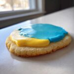 Close-up of a baked sugar cookie topped with bright yellow and blue icing using the sugar cookie icing recipe.