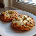 Two freshly baked pizza bagels topped with melted, bubbly cheese and herbs sitting on a white plate.