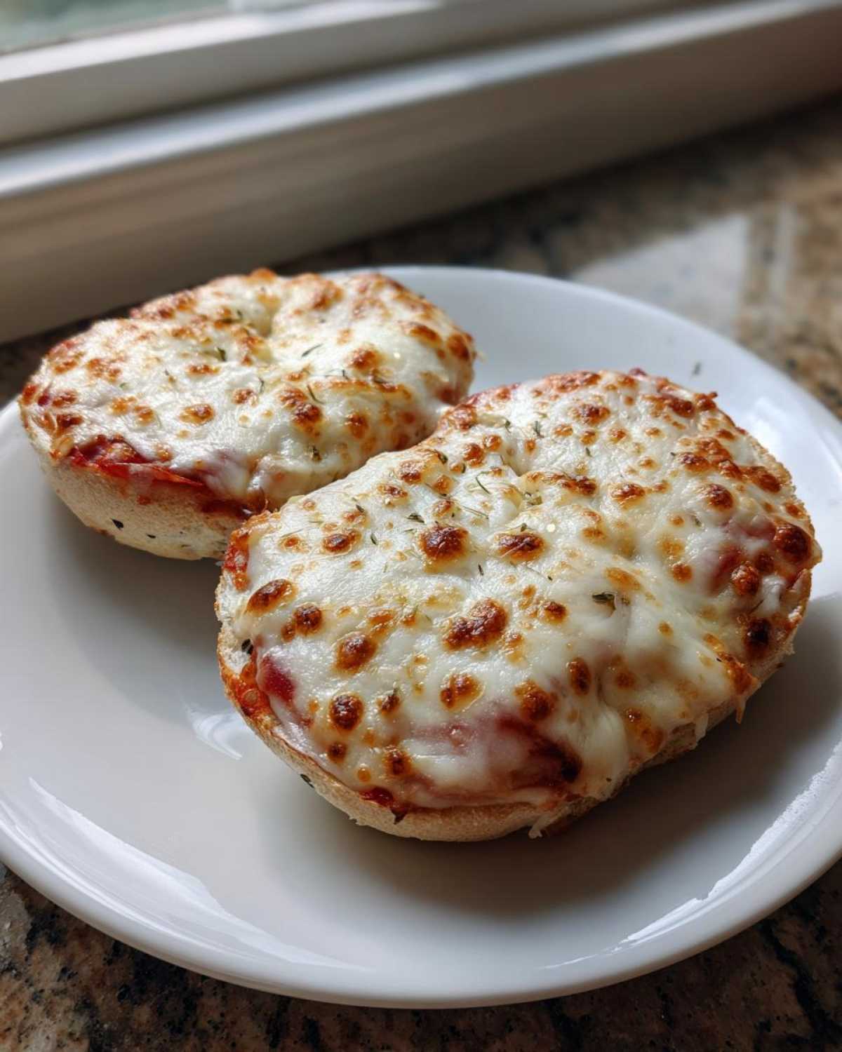 Two freshly baked pizza bagels topped with melted, bubbly mozzarella cheese and herbs, served on a white plate.
